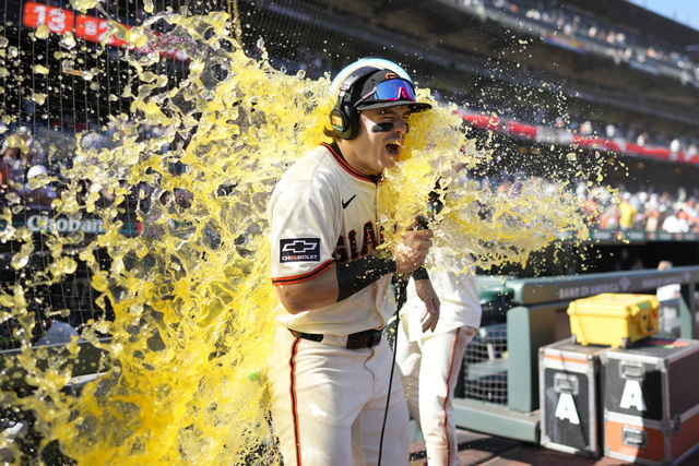 A baseball player in a white uniform and helmet is hit with a splash of yellow liquid while holding a microphone near the dugout during a game.